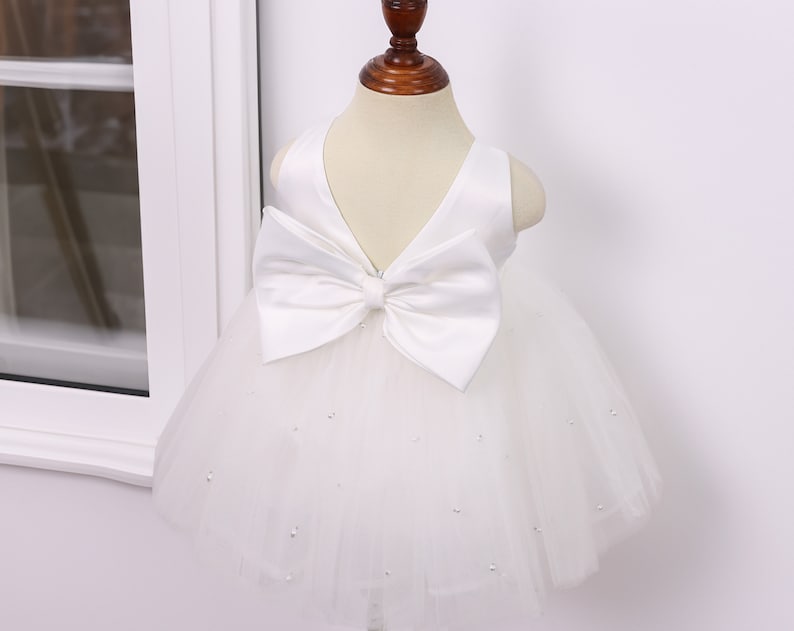 May include: White tulle baby dress with a large white satin bow on the bodice. The dress is decorated with small white beads.