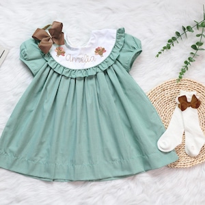 May include: A green and white gingham dress with a ruffled neckline and short sleeves. The dress has a white collar with embroidered turkeys and the name "Amelia" on the front.