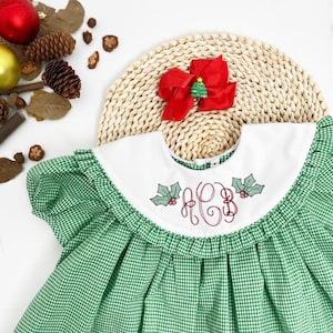 May include: A green and white gingham dress with a white collar featuring embroidered holly and initials. A red bow with a small Christmas tree is placed on a woven mat. Festive holiday attire.