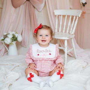 May include: A baby wearing a pink gingham romper with a white bib embroidered with the name "Everlee" and red hearts. The baby has a red bow in their hair and red bows on their white socks. A white wooden chair and a vase of white flowers are in the background.