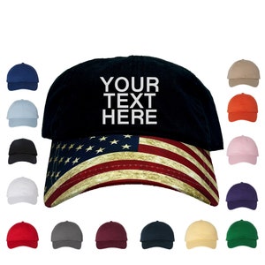 May include: A black baseball cap with a distressed American flag design on the brim. The front of the cap has white text that says "YOUR TEXT HERE". The cap is available in a variety of colors.