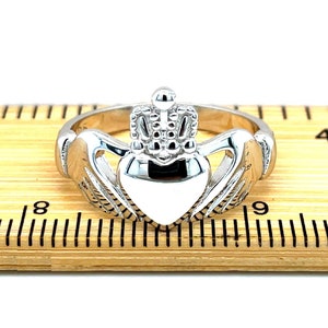 May include: A silver Claddagh ring featuring a heart, crown, and two hands. The ring is displayed on a wooden ruler, measuring approximately 8.5 inches in diameter.