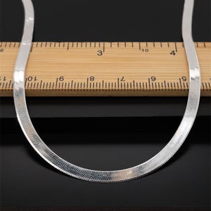 Op de afbeelding: Een zilveren visgraat ketting. De ketting is plat en flexibel, met een geweven patroon. De ketting wordt getoond op een houten liniaal met inch markeringen tegen een zwarte achtergrond. Het is een sieraad.