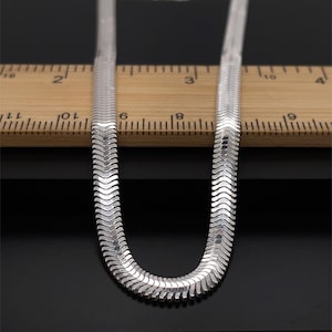 May include: A silver herringbone chain necklace. The flat, flexible chain has a woven pattern. It's shown against a wooden ruler and a black background. The necklace is approximately 40 cm long.