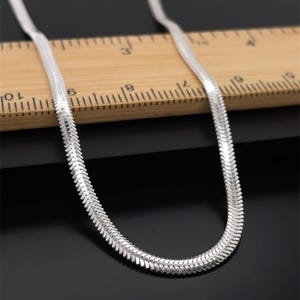 May include: A silver herringbone chain necklace. The necklace is on a wooden ruler with black markings. The chain has a flat, flexible design, and the links are closely connected, creating a smooth, reflective surface.
