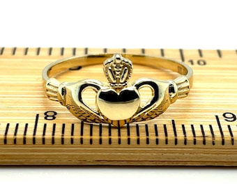 May include: A gold-colored Claddagh ring with a heart, crown, and hands. The ring is on a wooden ruler.