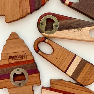 May include: Five wooden bottle openers with a variety of wood grain patterns and colors. Each opener has a brass bottle opener and the text "Beckloff Woodworks" is engraved on the wood.