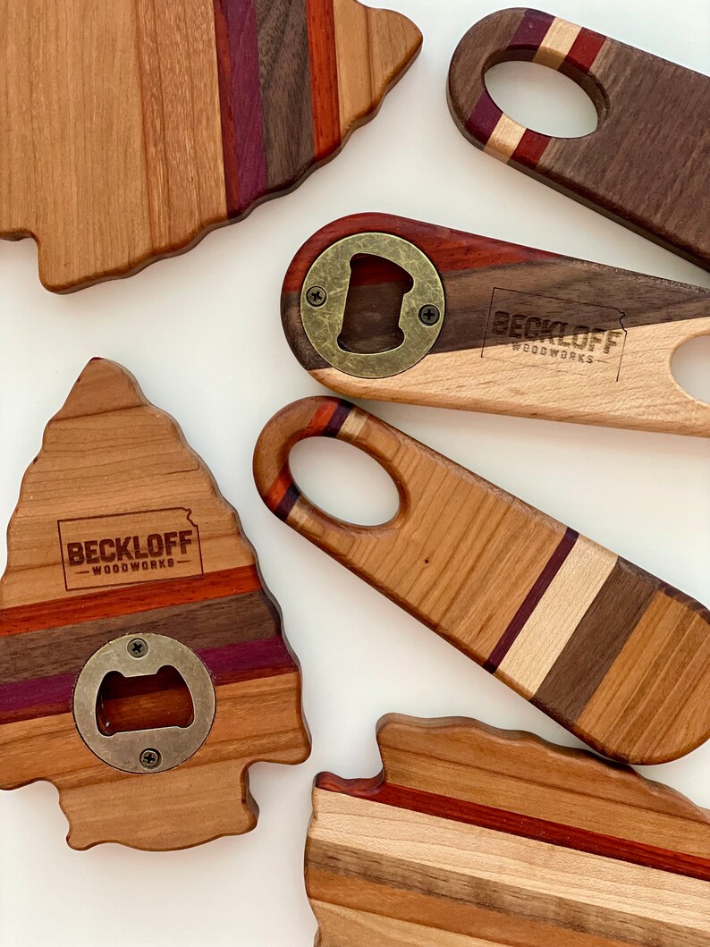 May include: Five wooden bottle openers with a variety of wood grain patterns and colors. Each opener has a brass bottle opener and the text "Beckloff Woodworks" is engraved on the wood.