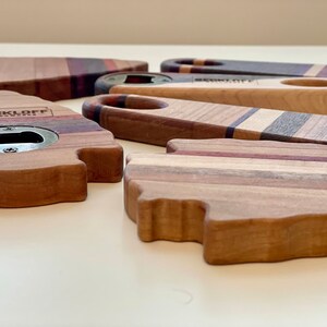 May include: A collection of five wooden bottle openers with different designs. The openers are made of various types of wood, including maple, walnut, and cherry. The openers have a metal bottle opener built into the wood. One opener has the text "BECKLOFF WOODWORKS" engraved on it.