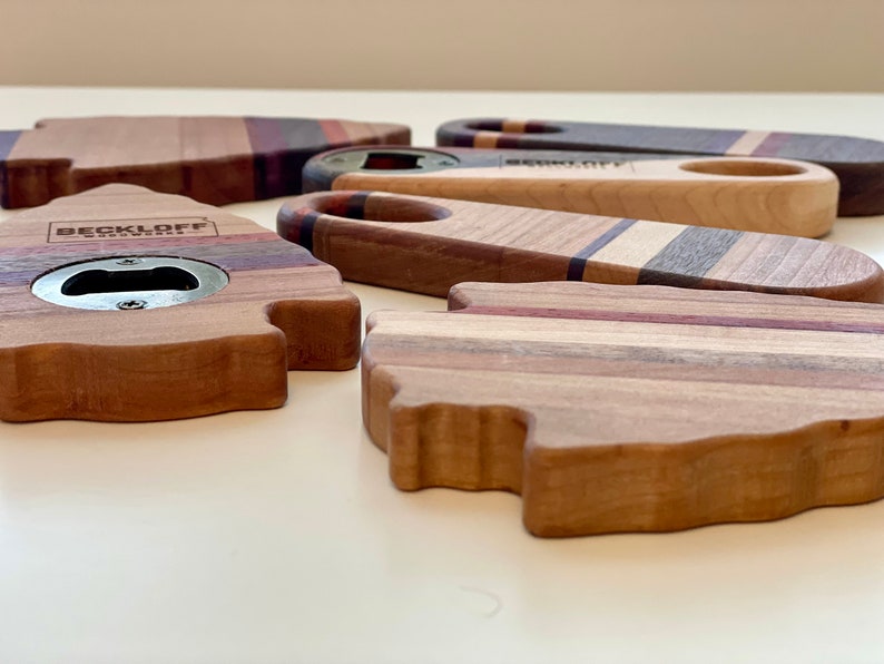 May include: A collection of five wooden bottle openers with different designs. The openers are made of various types of wood, including maple, walnut, and cherry. The openers have a metal bottle opener built into the wood. One opener has the text "BECKLOFF WOODWORKS" engraved on it.