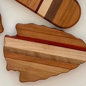 May include: A wooden cutting board shaped like an arrowhead with a multi-colored wood grain pattern. The board has a natural wood finish and is made of different types of wood.