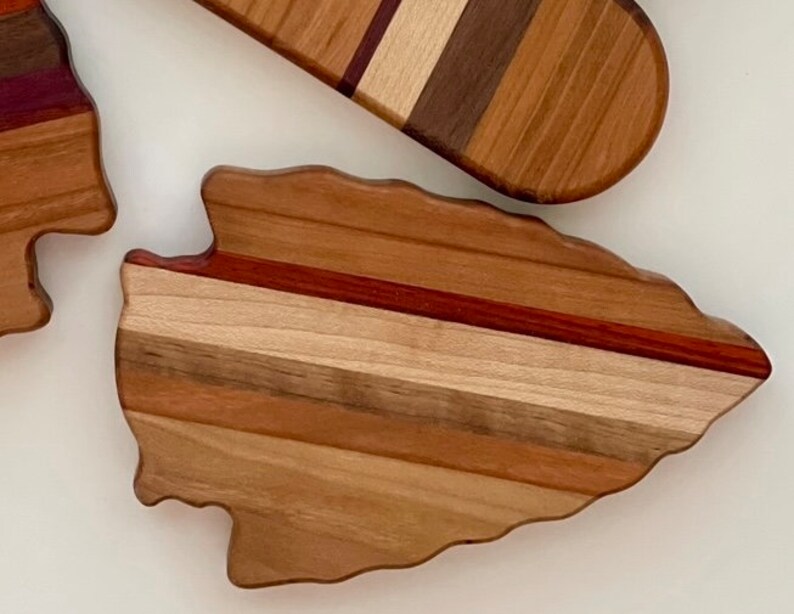 May include: A wooden cutting board shaped like an arrowhead with a multi-colored wood grain pattern. The board has a natural wood finish and is made of different types of wood.