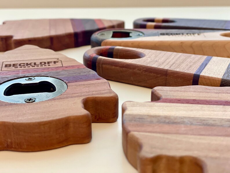 May include: A set of four wooden bottle openers, each with a unique shape and design. The openers are made from different types of wood, including maple, walnut, and cherry. The openers are all finished with a clear coat to protect the wood. The openers are all engraved with the words "Beckloff Woodworks".