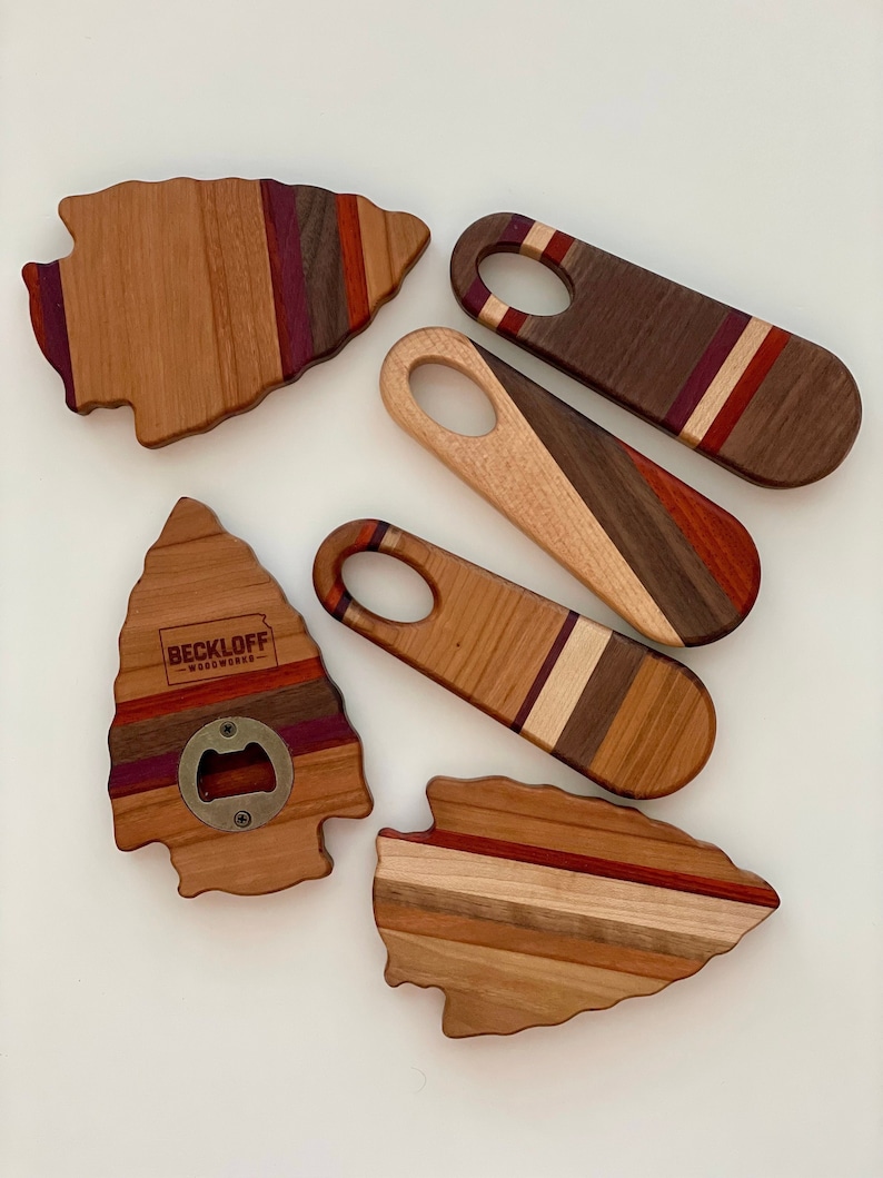 May include: Five wooden bottle openers in the shape of arrowheads. The bottle openers are made of different types of wood and have a variety of colors, including red, brown, and white. One bottle opener has the text "BECKLOFF WOODWORKS" engraved on it.