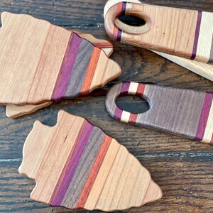 May include: Three wooden arrowhead-shaped cutting boards with a multi-colored striped design. The boards are made of different types of wood, including walnut, maple, and cherry. The boards are arranged in a staggered pattern on a wooden surface.
