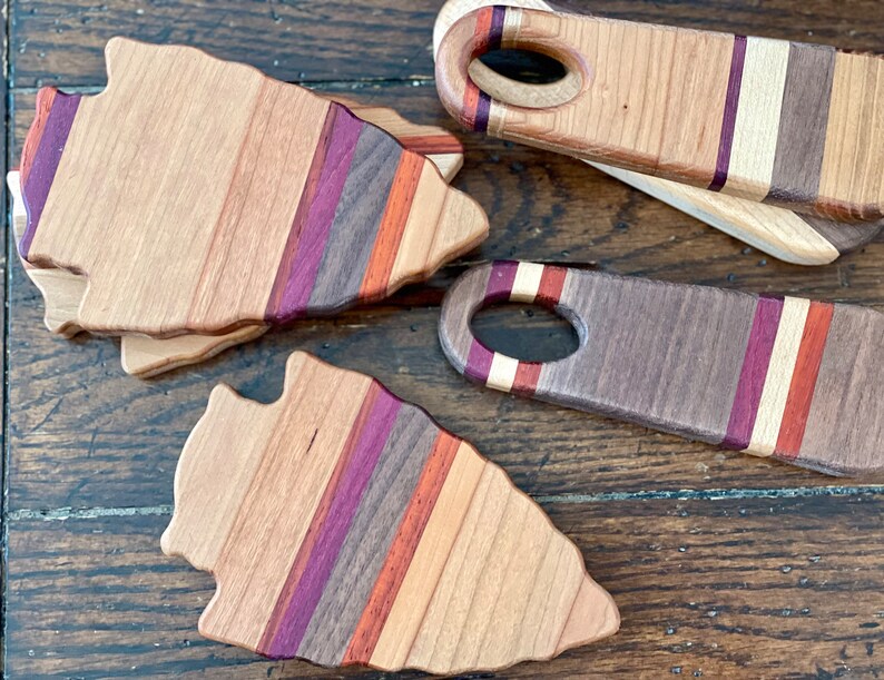 May include: Three wooden arrowhead-shaped cutting boards with a multi-colored striped design. The boards are made of different types of wood, including walnut, maple, and cherry. The boards are arranged in a staggered pattern on a wooden surface.