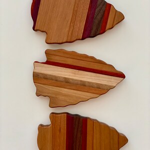 May include: Three wooden arrowhead-shaped cutting boards with different wood grain patterns. The boards are made of various shades of brown, red, and purple wood.