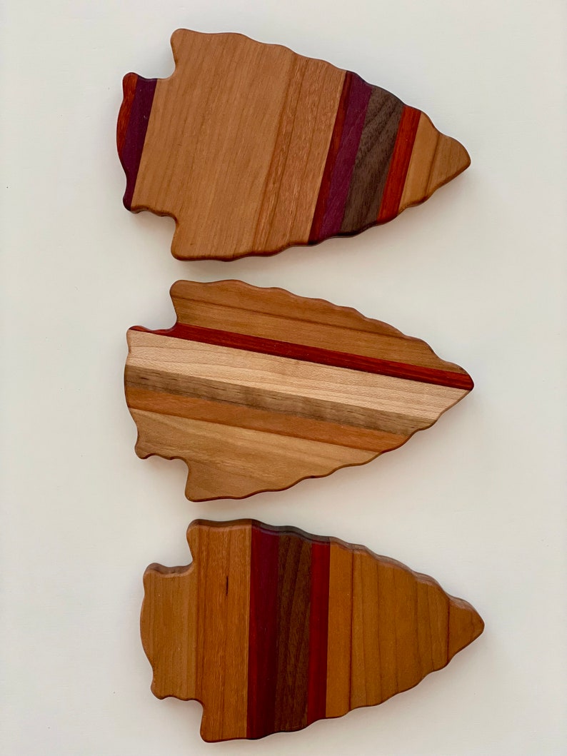 May include: Three wooden arrowhead-shaped cutting boards with different wood grain patterns. The boards are made of various shades of brown, red, and purple wood.