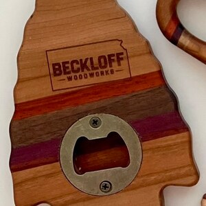May include: A wooden bottle opener shaped like an arrowhead with a metal bottle opener in the center. The wood is a mix of light and dark brown with purple and red stripes. The text "BECKLOFF - WOODWORKS" is engraved on the top of the opener.