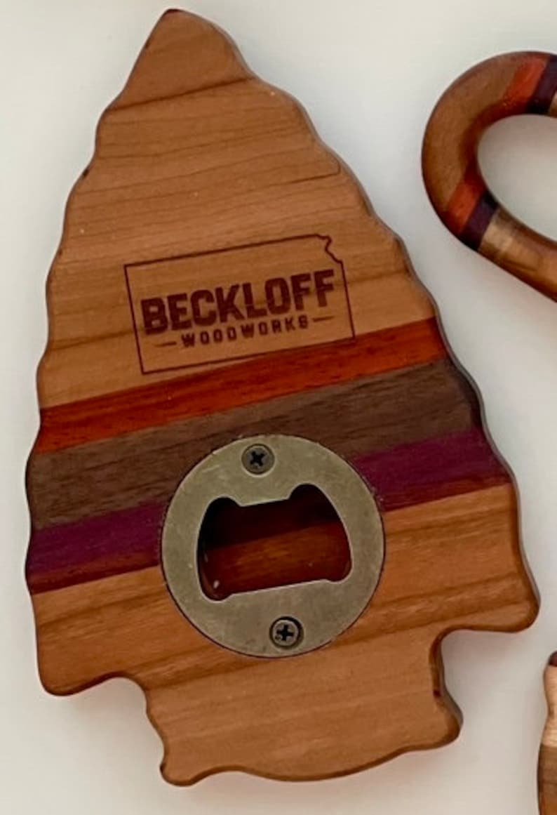 May include: A wooden bottle opener shaped like an arrowhead with a metal bottle opener in the center. The wood is a mix of light and dark brown with purple and red stripes. The text "BECKLOFF - WOODWORKS" is engraved on the top of the opener.