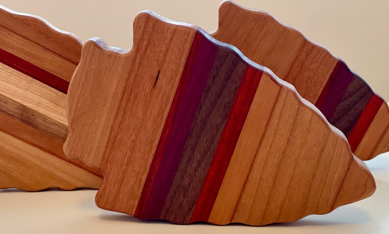 May include: Three wooden arrowhead-shaped cutting boards with a layered design of different wood types, including cherry, maple, walnut, and purpleheart. The boards are arranged in a staggered formation.
