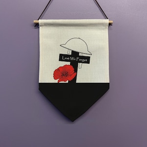 Pode incluir: Uma faixa de lona branca com uma cruz preta, uma papoula vermelha e um capacete preto e branco. A cruz tem as palavras "Lest We Forget" escritas nela.