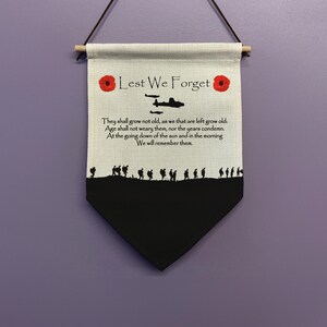 May include: A white canvas banner with the words "Lest We Forget" printed in black. The banner also features a silhouette of a plane and a line of soldiers marching. The banner is decorated with two red poppy flowers.