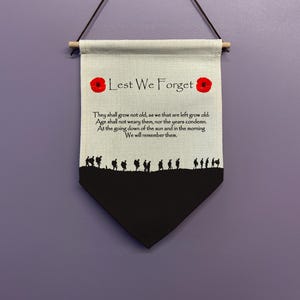 May include: A beige canvas banner with the words "Lest We Forget" in black lettering and two red poppy flowers. The banner also features a black silhouette of a line of soldiers marching across a black field.