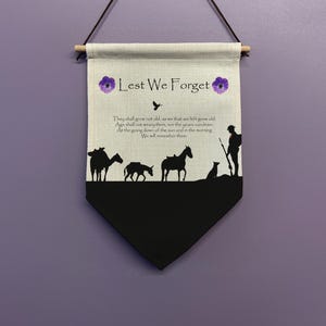 Puede incluir: Una pared colgante de lona beige con las palabras "Lest We Forget" en texto negro. La pared colgante presenta una silueta de un soldado, un camello, un caballo y un perro. El fondo es una pared morada.