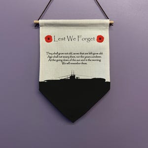 May include: A white canvas banner with the words "Lest We Forget" printed in black. The banner has two red poppy flowers on either side of the text. The banner also has a black silhouette of a submarine at the bottom. The text below the submarine reads: "They shall grow not old, as we that are left grow old: Age shall not weary them, nor the years condemn. At the going down of the sun and in the morning We will remember them."