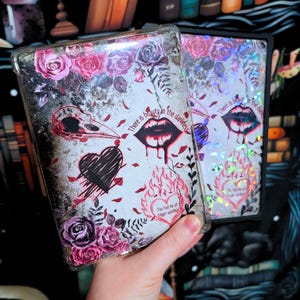 May include: Two phone cases with a gothic aesthetic. One case features a black and white heart, a skull, and a red heart with flames. The other case is iridescent and features a heart with flames. Both cases have the text "There is beauty in the darkness" and "You had me at trigger warning".