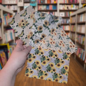 May include: Three decorative paper sheets with floral patterns. The top sheet features a butterfly and floral design in shades of brown, white, and green. The middle sheet has a vintage newspaper print with a floral design in shades of brown, white, and green. The bottom sheet has a floral design in shades of brown, white, and green.