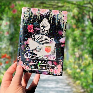 May include: A black and white illustration of a skeleton reading a book with a peach on the cover. The illustration is surrounded by pink flowers and a glittery background. The text "The Reader" is at the bottom of the image.