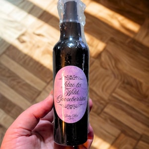 May include: A dark purple body mist bottle with a label that reads "Lilac & Wild Gooseberries." The label is oval-shaped with a light purple background and decorative floral elements. The bottle is held in a hand.