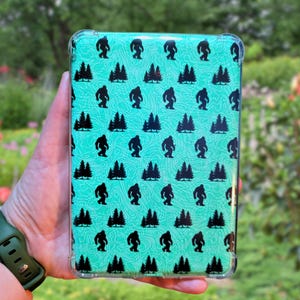 May include: A clear phone case with a teal background and a repeating pattern of black silhouettes of Bigfoot and pine trees.