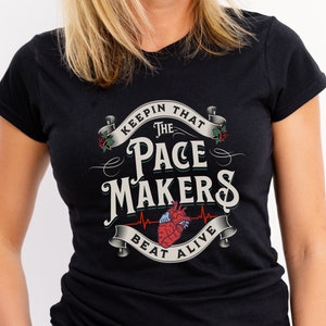 Puede incluir: Camiseta negra con un diseño gráfico blanco que presenta un corazón con una línea de latido del corazón y el texto "Keepin' That The Pace Makers Beat Alive".