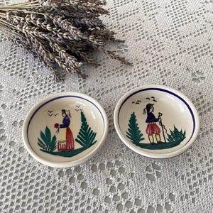 Può includere: Due piccole ciotole rotonde in ceramica con disegni di arte popolare dipinti a mano. Una ciotola raffigura una donna con fiori, l'altra una persona con un bastone. Entrambe hanno un bordo blu e accenti verdi e rossi. Lavanda secca sullo sfondo.