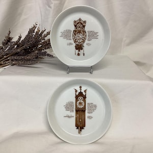 May include: Two white ceramic plates with brown clock designs. The plates have a raised rim and are decorated with a clock face and a clock tower. The clock tower is surrounded by text in a brown font.