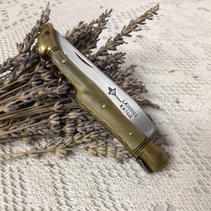May include: A folding pocket knife with a metallic handle and a silver blade. The blade features the engraved text LAGUIOLE EXTRA and a small symbol of a cross. The knife is positioned diagonally across a bundle of dried lavender stems.