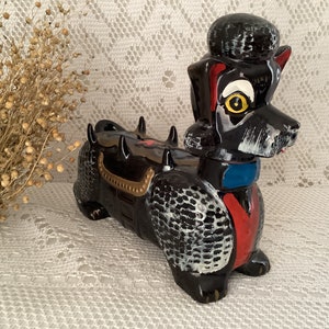 May include: A black ceramic poodle figurine with white and gold accents. The poodle has a blue and red collar and a yellow eye.