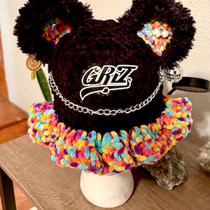 May include: A black crocheted bucket hat with two large pom-poms on top, featuring a vibrant multicolored ruffled brim. The hat displays the text "GRiZ" in white, a silver chain across the front, and small charms including a rainbow and lollipop.