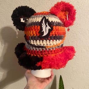 May include: A handmade, colorful crochet hat with black, orange, red, gray, and white stripes. The hat features two fluffy ear-like pom-poms, a patch with a white and black design, and decorative chains. Small charms and a lollipop are attached.