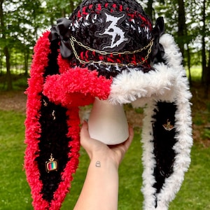 May include: A black, red, and white crocheted hat with long, plush ear flaps. The hat features a white embroidered patch, a gold-tone chain, and black ribbon bows. The long ear flaps are adorned with small metal charms, including a television and a moth.