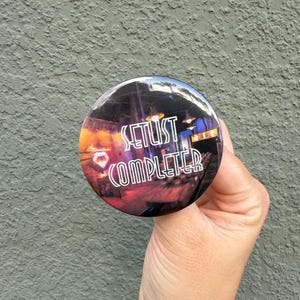 May include: A circular pin-back button featuring a photographic image of a dimly lit indoor venue with warm orange and cool blue lighting. The text SETLIST COMPLETER is printed in a bold, white, stylized sans-serif font across the center.