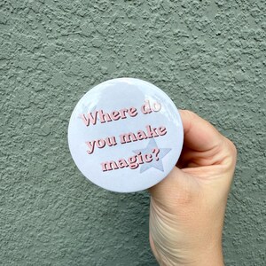 May include: A circular pin-back button with a white background featuring the text "Where do you make magic?" in pink letters. The design includes a light gray star graphic behind the text, creating a simple and clear visual display on the button.