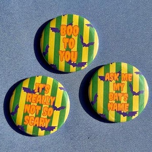 May include: Three circular pin-back buttons featuring a vertical green and yellow striped pattern with purple bat silhouettes. The buttons display orange text reading BOO TO YOU, IT'S REALLY NOT SO SCARY, and ASK ME MY BAT'S NAME in a bold, playful font.