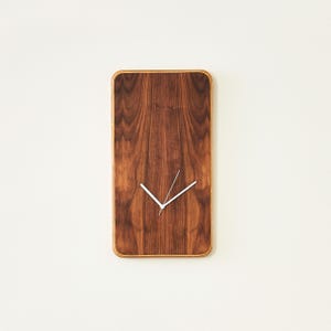 May include: A rectangular wall clock with a dark brown wood grain face and rounded corners. The clock features two thin, white hands positioned in the center, indicating the time without any numbers or hour markers on the wooden surface.
