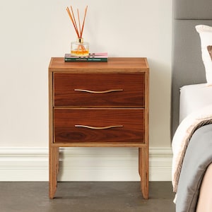 May include: A wooden nightstand with two drawers featuring horizontal wood grain patterns and thin, wavy gold-toned metal handles. The unit has a layered plywood edge design and stands on four legs. A glass reed diffuser and a book sit on top.