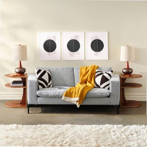 May include: A modern living room setup featuring a gray sofa with black and white geometric pillows and a mustard yellow throw blanket. Two wooden side tables with lamps flank the sofa. Three framed abstract art pieces hang above the sofa, and a white shag rug is on the floor.