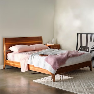 May include: A wooden bed frame with a headboard, featuring a light pink sheet set, white quilted comforter, and a pink throw blanket. The bed is in a well-lit room with a small wooden nightstand and a decorative rug.